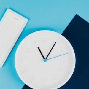 Photo by www.kaboompics.com Stylish flat lay with a white clock and notepad on blue paper background.